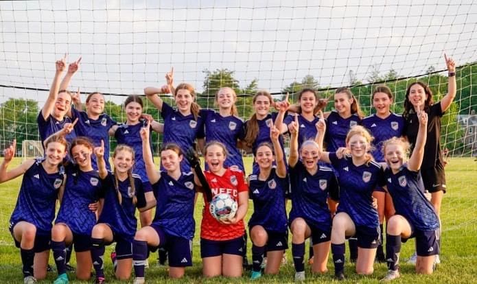 A joyful girls' soccer team posing in front of a goal with raised index fingers.