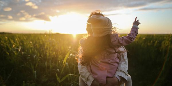 Person holding a child pointing at the sunset in a field.
