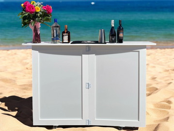 A white beach bar cart with drinks and flowers against ocean backdrop.