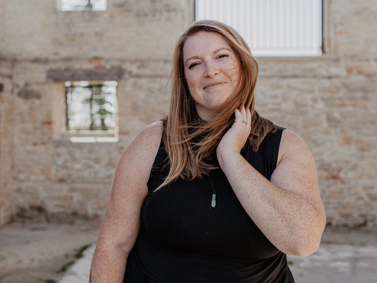 Smiling woman in black dress standing against a rustic stone wall.