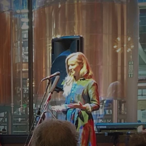 Woman speaking at a microphone in a modern indoor setting with large copper tanks.