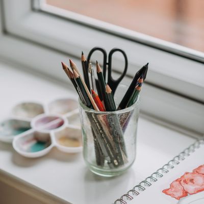 A glass jar holding pencils and scissors on a windowsill with watercolor paints and artwork nearby.