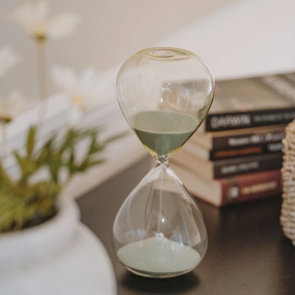 A glass hourglass with green sand on a dark surface near plants and books.