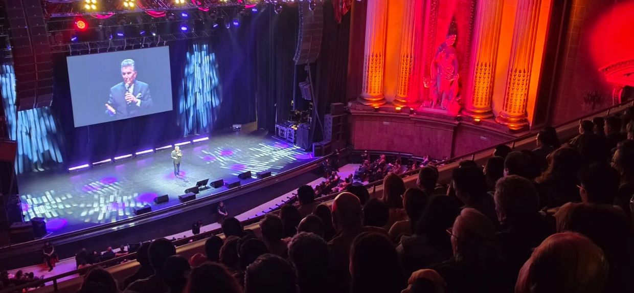 A speaker on stage in a large theater with an audience seated in balconies.