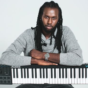 Man with dreadlocks leaning on a keyboard piano.