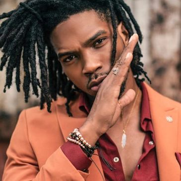 Man with dreadlocks wearing an orange blazer and red shirt, posing thoughtfully.