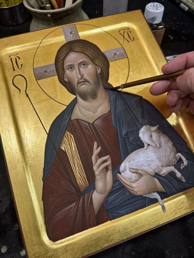 Hand painting a religious icon of Jesus holding a lamb on a gold background.