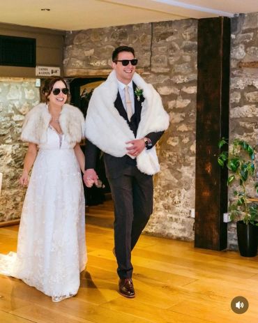A joyful bride and groom wearing sunglasses and fur wraps, holding hands indoors.