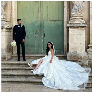 Bride in white gown sits on steps, groom in black suit holding bouquet.