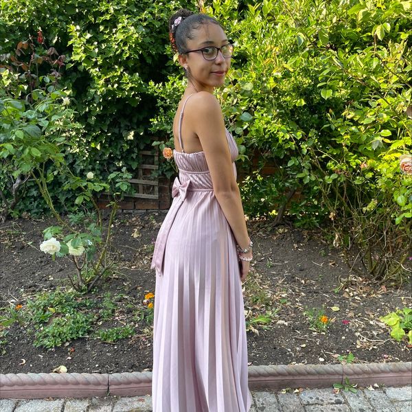 Young woman in a pink pleated dress stands in a garden with greenery.
