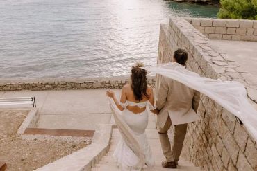 Newlyweds walking down stone steps by the water, bride's veil flowing in the breeze.