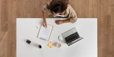 Person writing on clipboard next to an open laptop on a white desk.