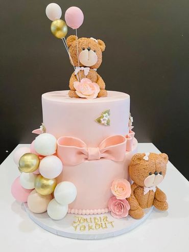 Two-tier pink cake with teddy bears, balloons, and floral decorations.