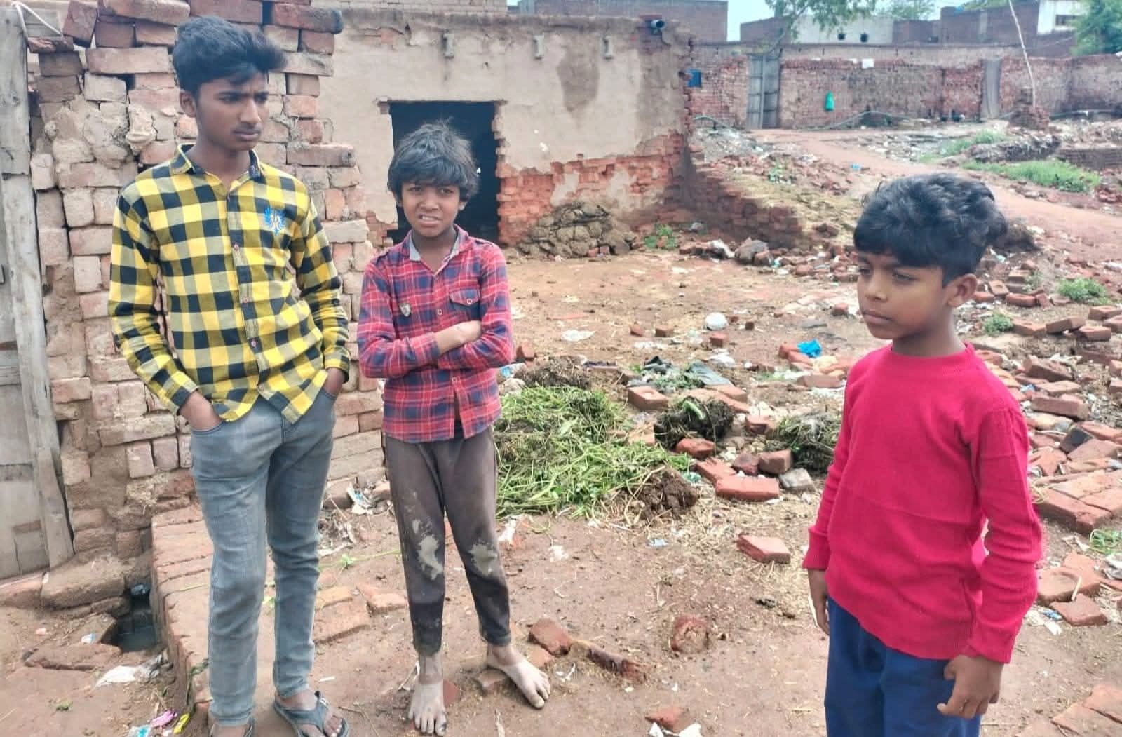 Three boys standing in a rustic, brick-walled outdoor area with dirt ground.