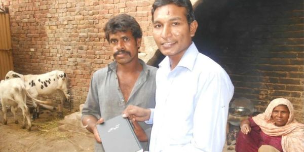 Two men, one holding a laptop, in a rustic courtyard with cows and a woman sitting.