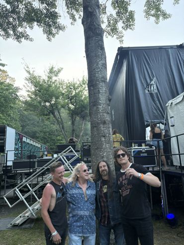 Four men posing casually outdoors near a stage setup with trees in the background.