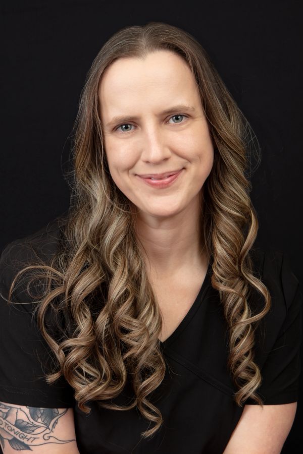 Smiling woman with long curly hair and a tattoo on her arm against a black background.