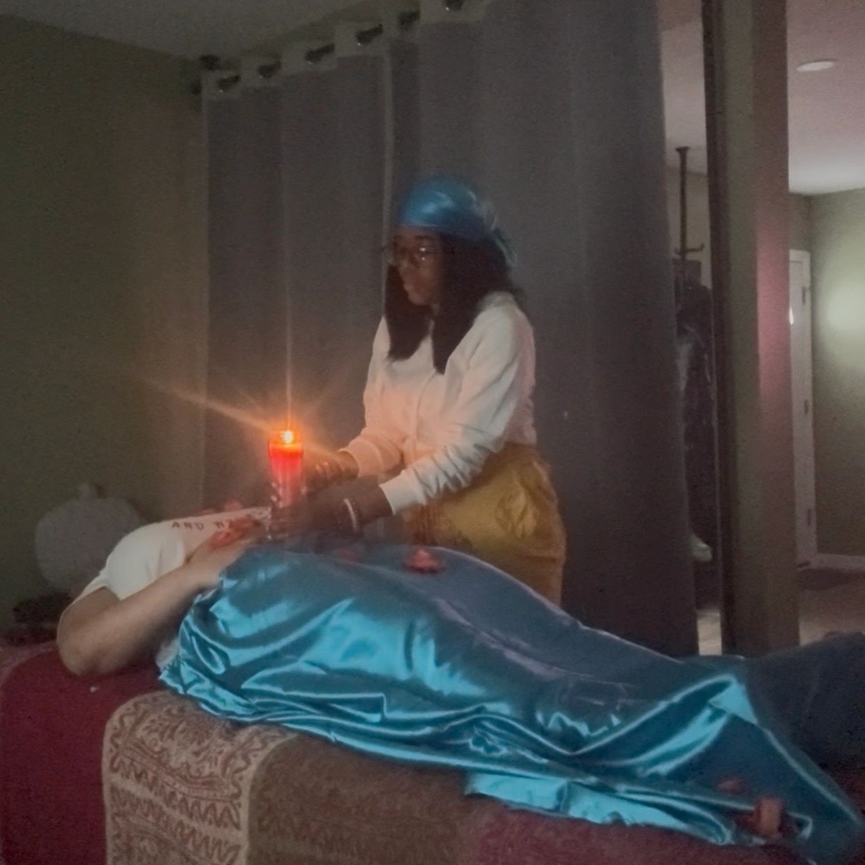 A woman holding a lit candle over another person covered in a blue satin cloth.