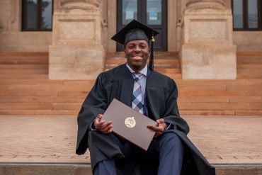 A happy graduate sitting on steps holding a diploma.