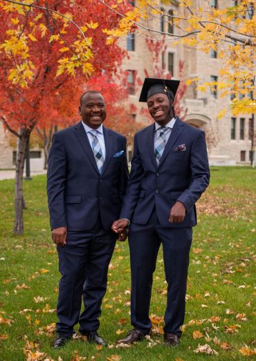Two men in matching suits, one wearing a graduation cap, smiling outdoors in autumn.