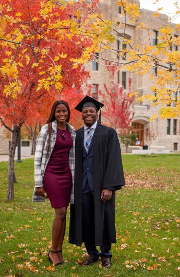 A graduate in cap and gown stands with a woman amidst colorful autumn trees.