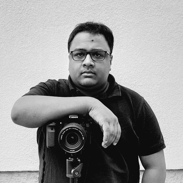 Man posing with a Canon camera on a tripod against a plain wall.