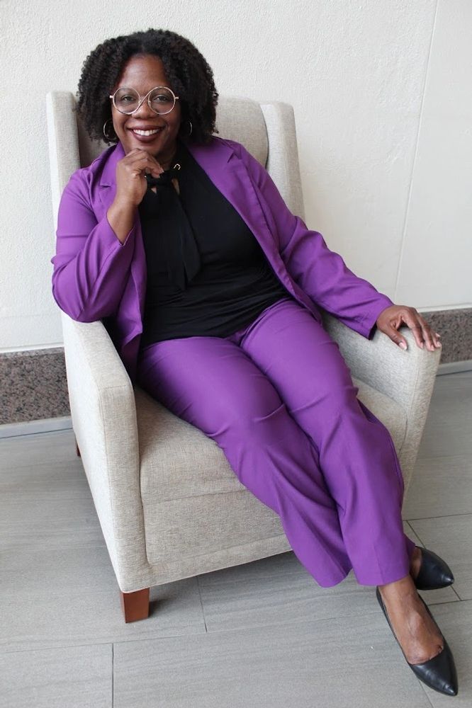 Smiling woman in purple suit sitting confidently in a beige armchair.