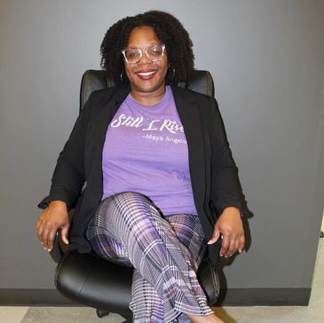Woman sitting confidently in a chair, wearing a purple 'Still I Rise' Maya Angelou t-shirt and plaid pants.