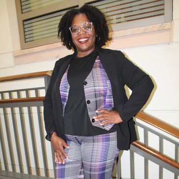 Woman in a purple plaid suit and black blazer smiling on stairs.
