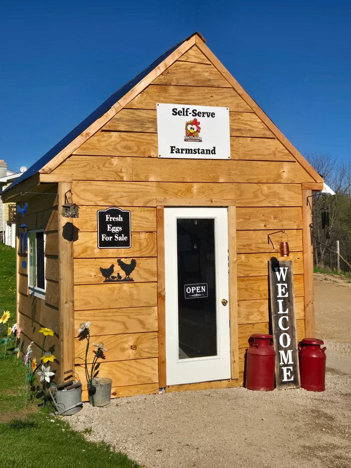 A rustic farm stand with a large rooster statue and signs advertising fresh eggs and homemade goods.