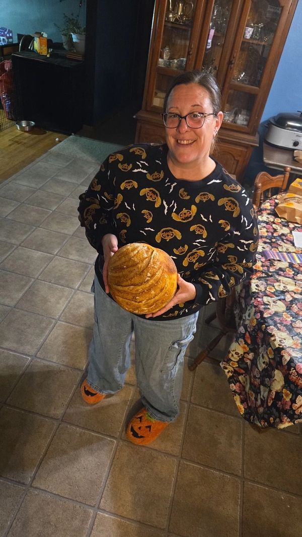 Woman smiling, holding freshly baked bread, wearing Halloween-themed clothes and slippers.