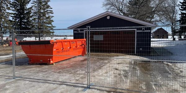 Orange dumpster behind a fenced area near a blue garage with snow on the ground.