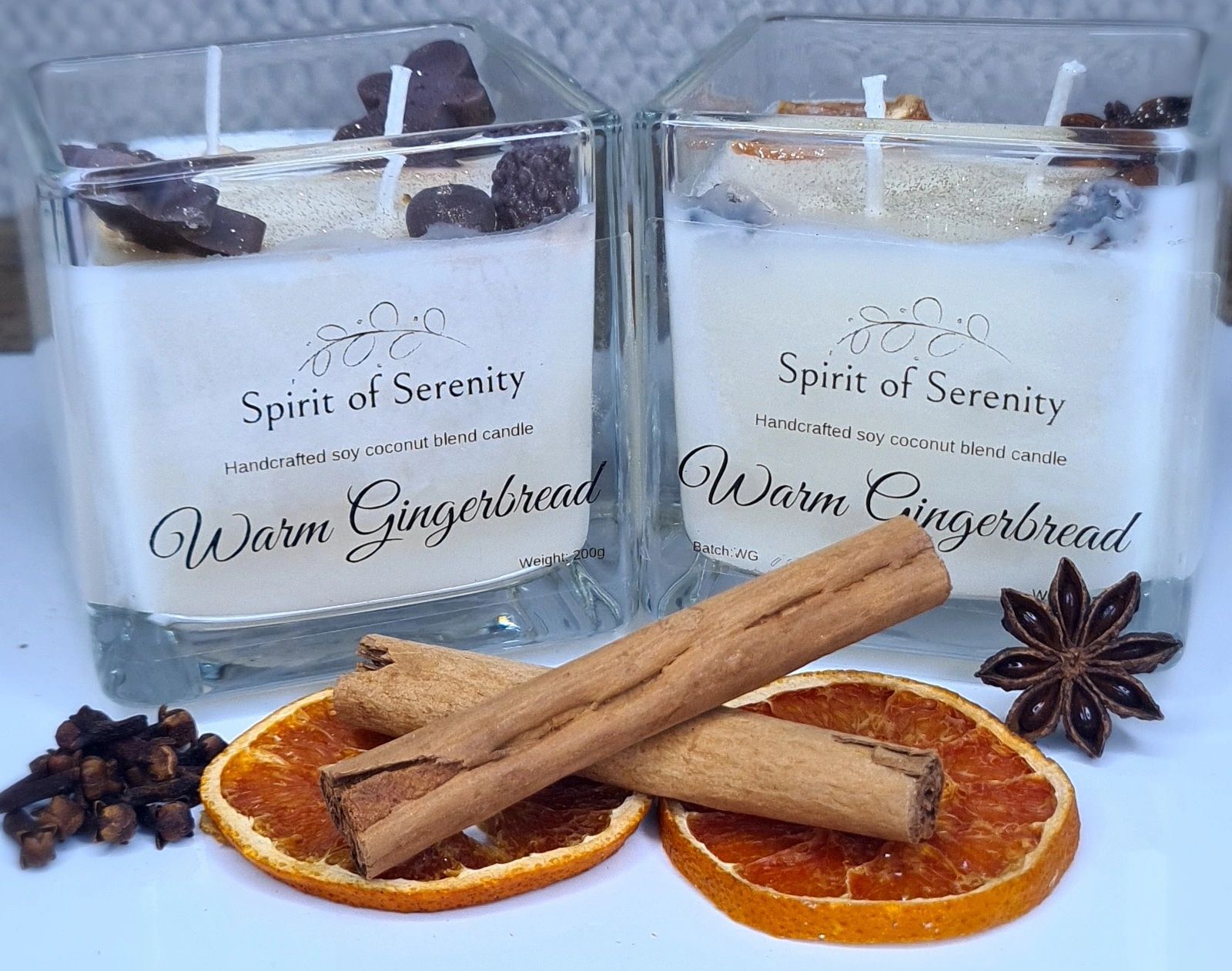 Photo of two white candles on a wooden base, next to two cinnamon sticks and two orange slices. 