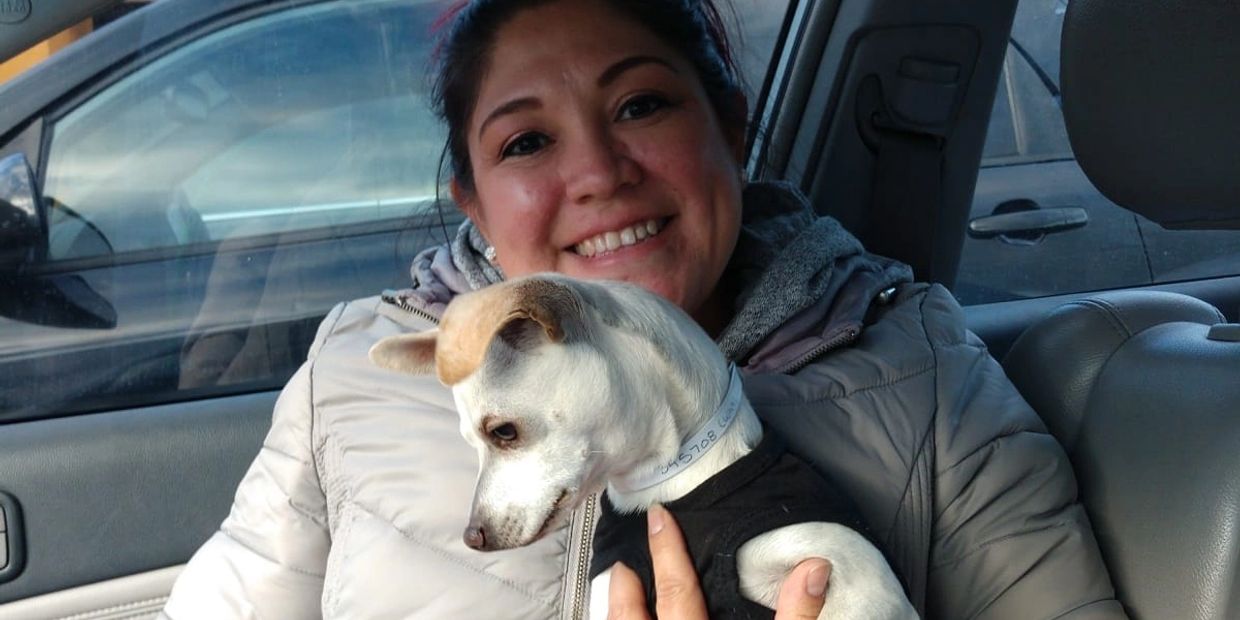 Woman smiling inside a car holding a small dog in a black outfit.