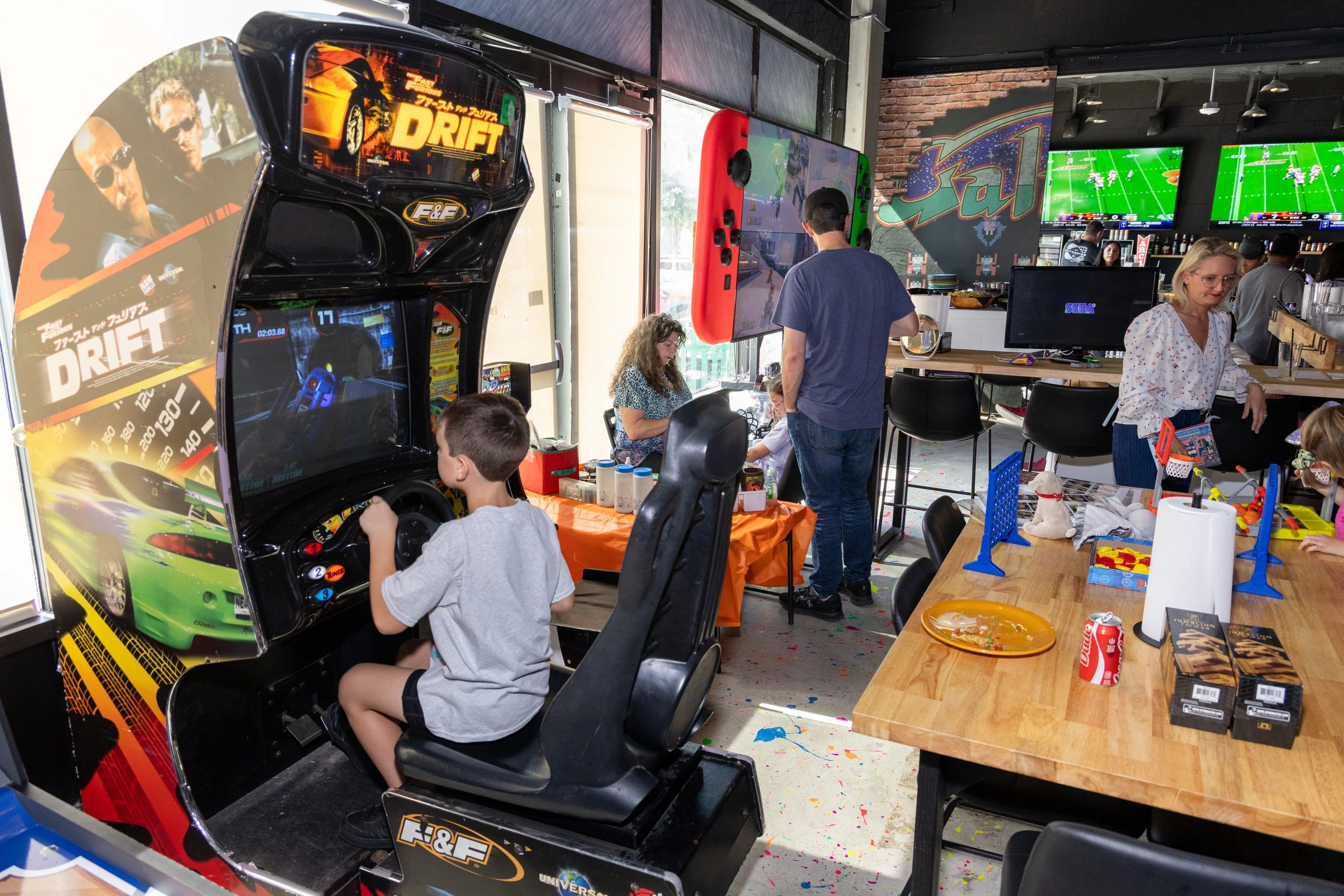 Kids and adults playing video games and arcade games in a lively gaming cafe.