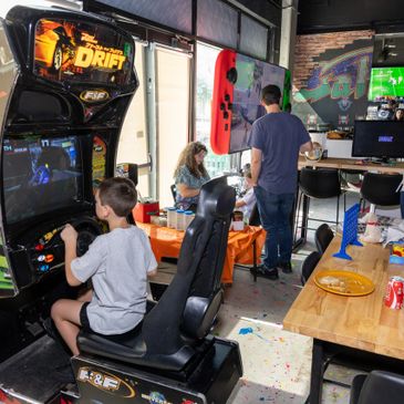 A boy plays arcade racing game while others socialize in a lively room.