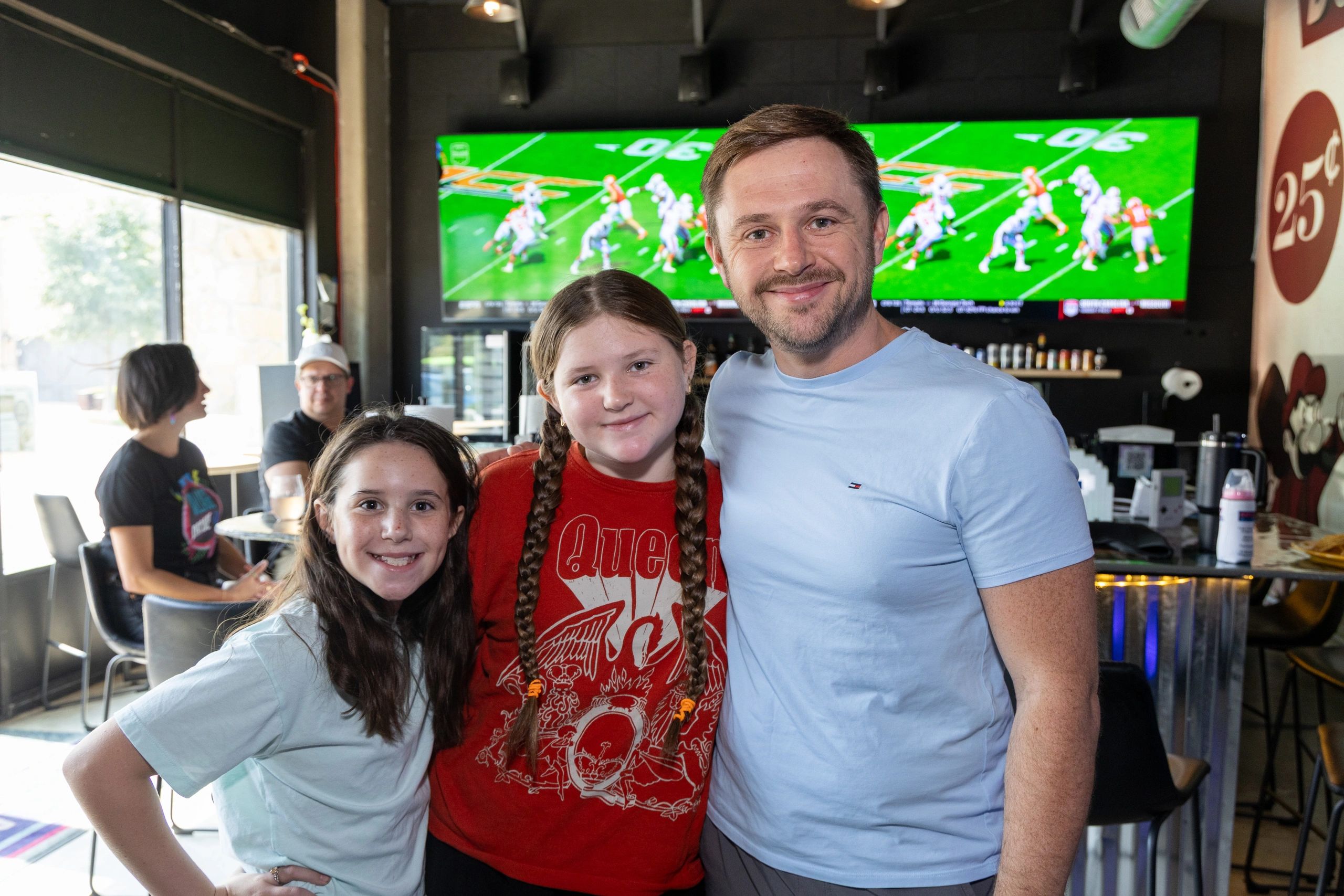 Smiling man and two girls posing in a sports bar with a football game on TV.