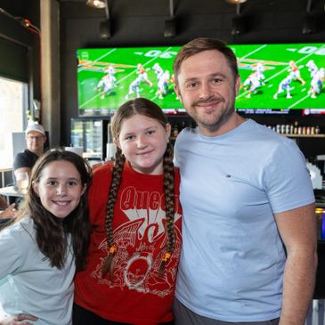 A man and two girls smiling indoors with a sports game on TV behind.