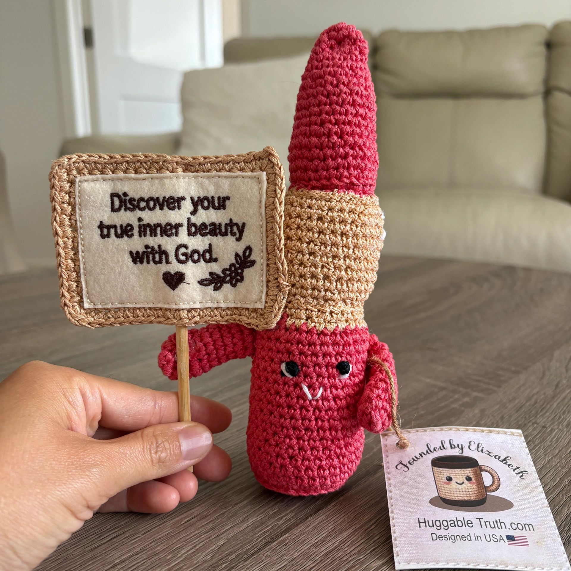Hand holding a crocheted red figure holding a sign with a religious quote.