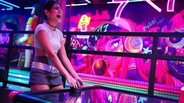 Young woman laughing while playing air hockey in a neon-lit arcade.