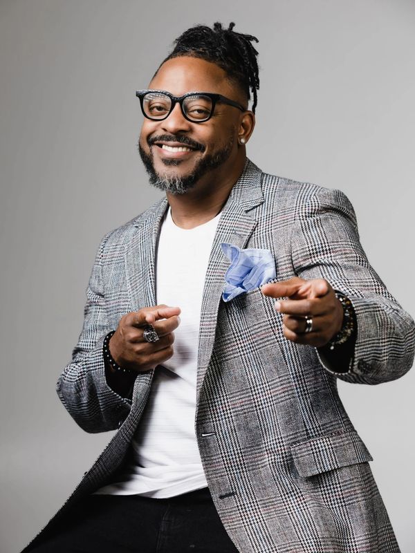 Smiling man in glasses and plaid blazer pointing playfully at the camera.