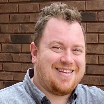 Smiling man with short curly hair and a beard against a brick wall.