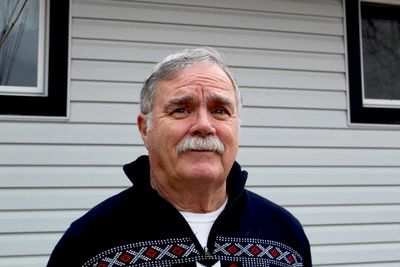 Older man with a mustache wearing a patterned sweater outdoors.