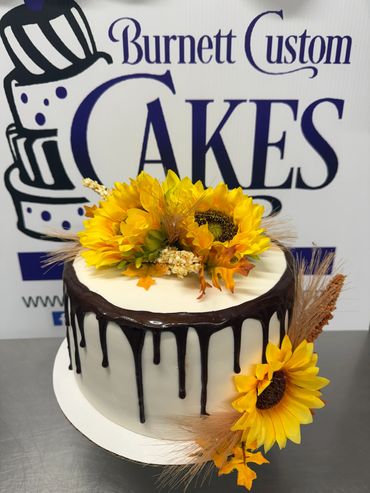 Elegant white cake with chocolate drip and sunflower decorations.