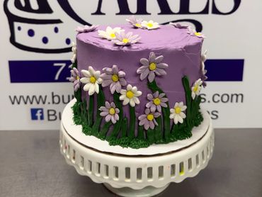 Purple cake decorated with white and purple daisies and green grass on a white stand.