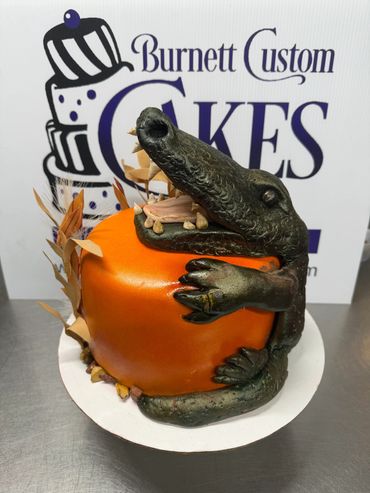 Cake shaped like an orange pumpkin being hugged by a realistic alligator figure.