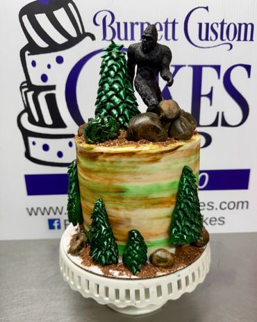 A nature-themed cake featuring a Bigfoot figure among green trees and rocks.