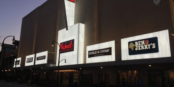 A row of illuminated storefront signs at dusk including Chipotle and Ben &amp; Jerry's.