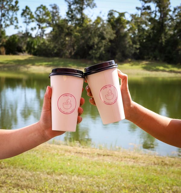 Two hands holding coffee cups by a lake on a sunny day.