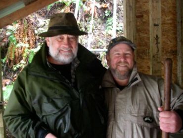 Two men smiling together inside a rustic cabin, one holding a wooden stick.